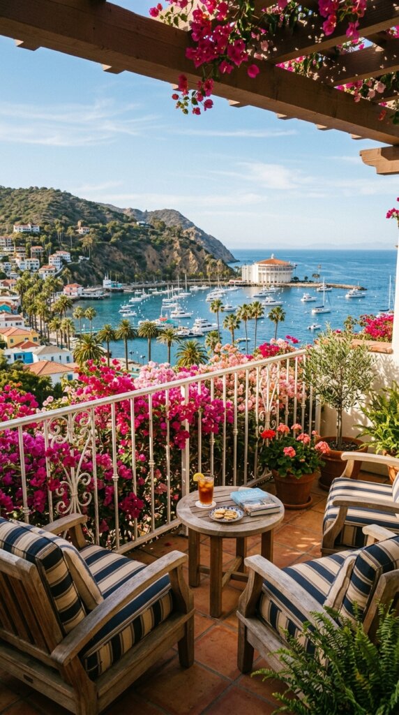 The view from a charming boutique hotel balcony or villa patio overlooking Avalon Harbor. Features white railings, colorful bougainvillea flowers, a cozy seating area, and a beautiful view of palm trees and ocean waters in the background. Warm, relaxing vacation atmosphere.